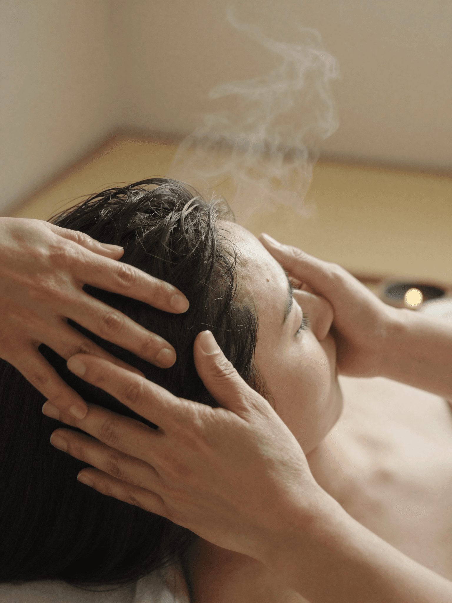 A therapist's hands performing a slow, precise scalp massage sequence, steam rising softly